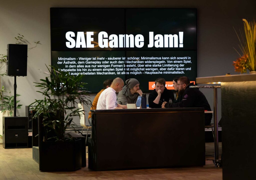 Personen sitzen gemeinsam an einem Tisch vor einem großen Bildschirm mit der Aufschirft Game-Jam.