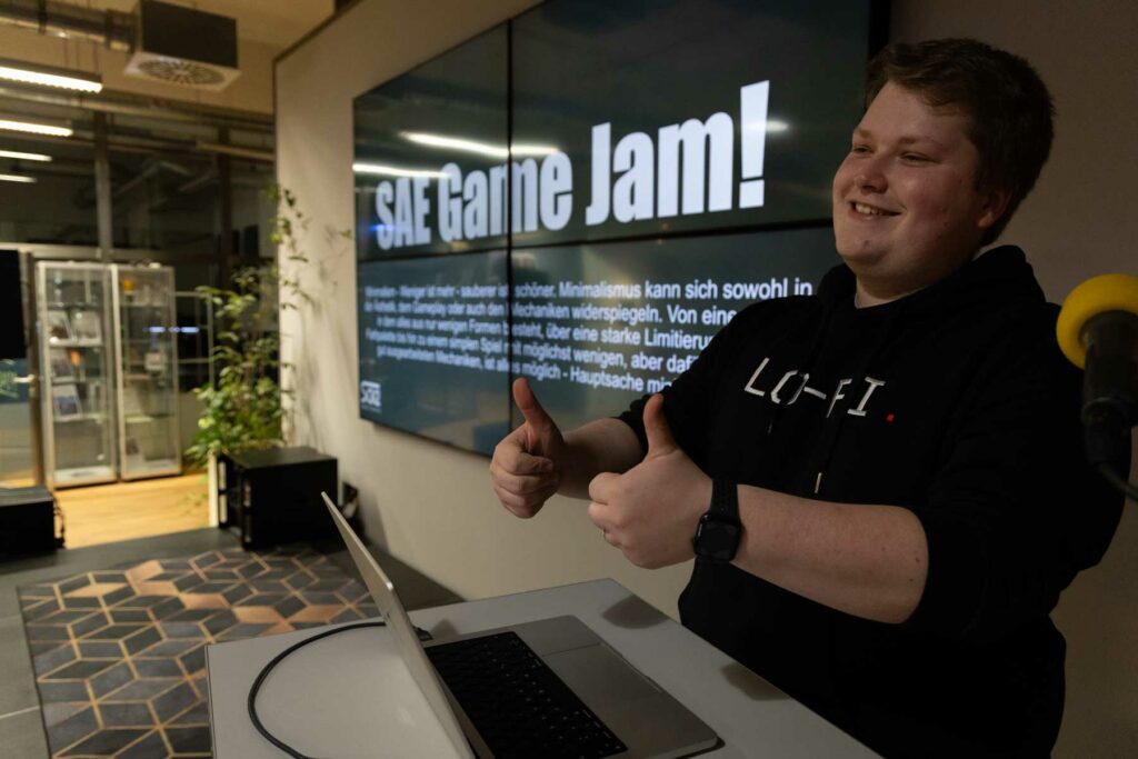 Student steht am Pult auf Bühne und zeigt die Daumen hoch.