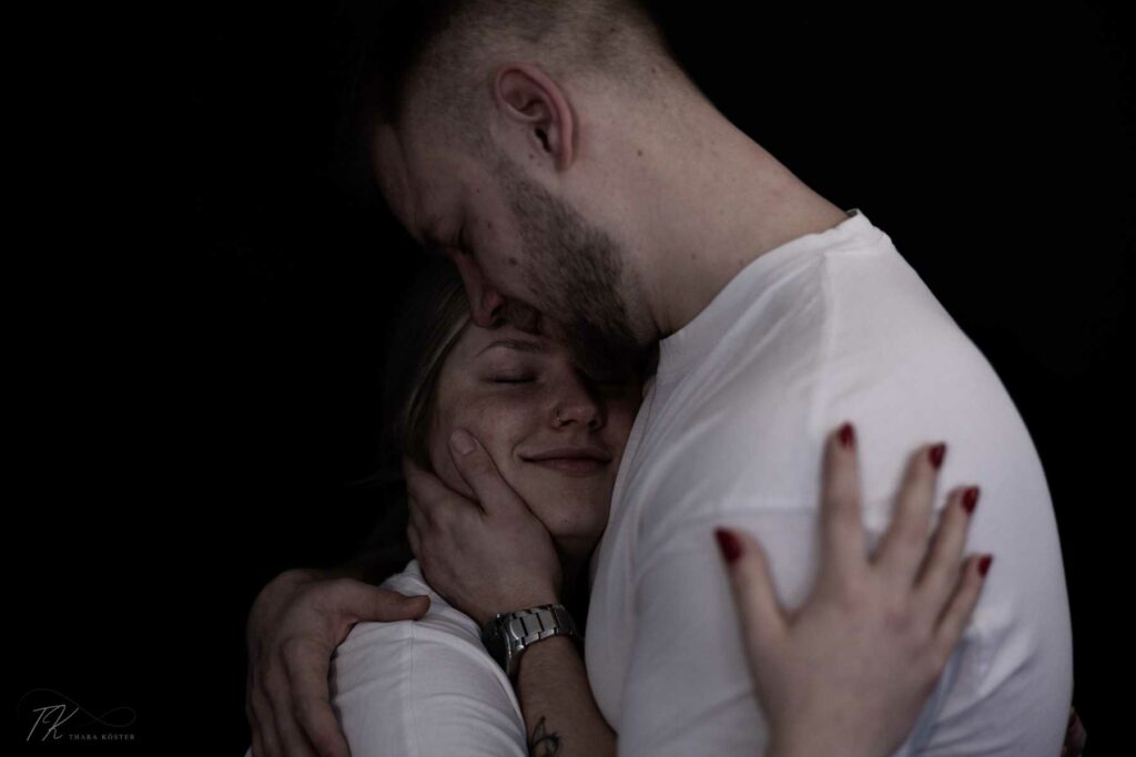 Zwei Menschen bei einem Paarshooting in heller Kleidung vor schwarzem Hintergrund die sich umarmen.