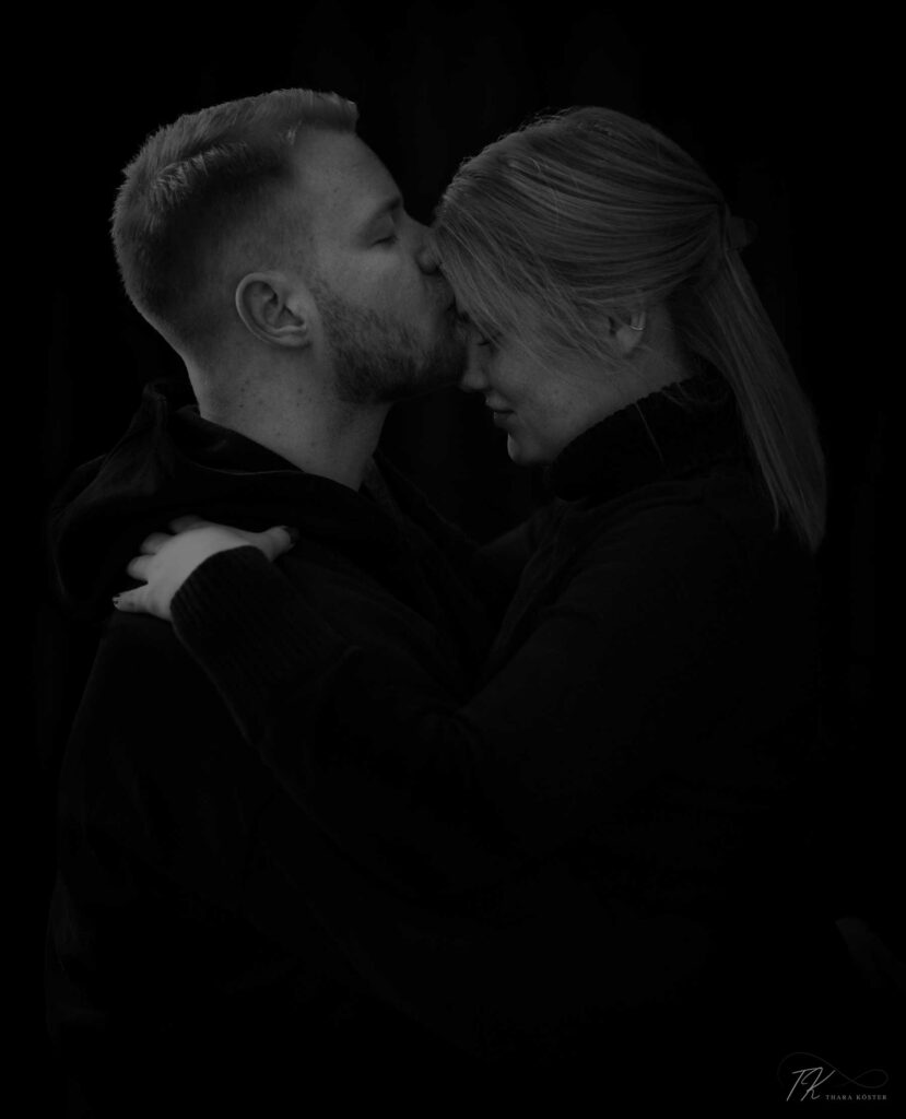 Zwei Menschen bei einem Paarshooting in dunkler Kleidung vor schwarzem Hintergrund sich umarmen und auf die Stirn küssen.