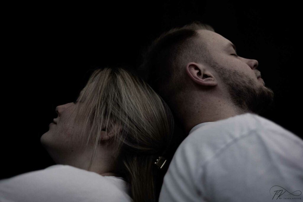 Zwei Menschen bei einem Paarshooting in heller Kleidung vor schwarzem Hintergrund die Rücken an Rücken gelehnt sind.