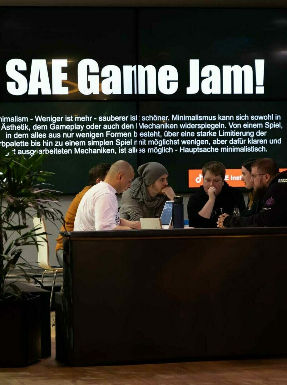 Studenten sitzen zusammen an einemt Tisch vor einem großen Bildschirm mit der Aufschrift SAE Game Jam